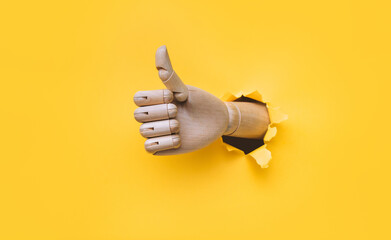 A wooden hand protrudes from a torn hole in yellow paper and shows a big thumbs up like gesture....
