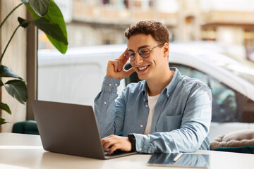 A cheerful young man with curly hair and glasses smiling at his laptop in a bright room with a plant
