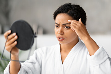 Concerned black woman looking in handheld mirror at pimple on her forehead