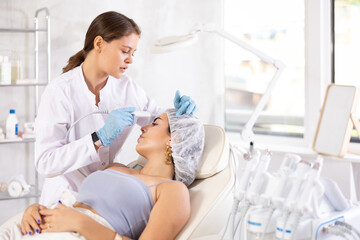 Professional female aesthetician administering hardware facial procedure on young woman in...