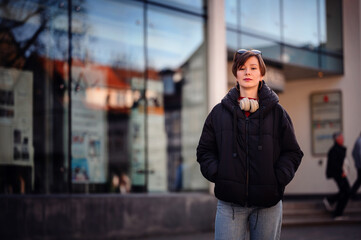 An urban woman stands confidently in front of a reflective glass building, headphones draped around...