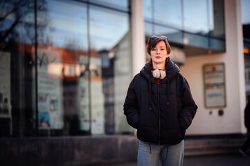 An urban woman stands confidently in front of a reflective glass building, headphones draped around...