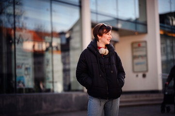 Fototapeta premium An urban woman stands confidently in front of a reflective glass building, headphones draped around her neck, blending city life and personal leisure