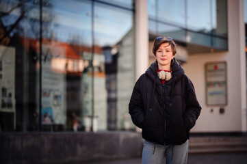 An urban woman stands confidently in front of a reflective glass building, headphones draped around...