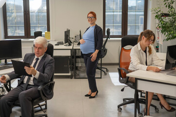 A happy pregnant woman stands in the middle of the office next to a Caucasian woman and an elderly man working at computers.