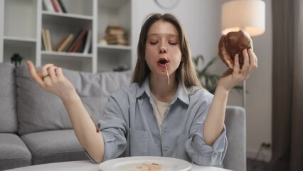 Portrait of funny happy young woman eating burger with great pleasure while ketchup flows down the hands at home Excited hungry female biting hamburger enjoying fast junk food indoors