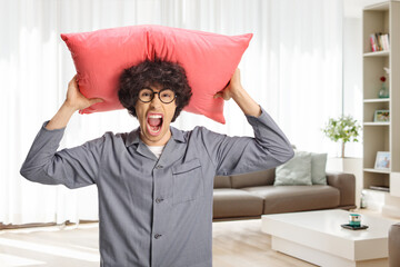 Angry young man in pajamas holding a pillow over head and screaming