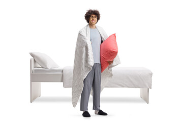 Young man in pajamas wrapped in a blanket standing next to a bed and holding a pillow