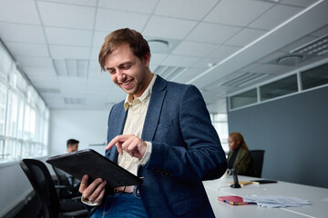 Happy young businessman in businesswear using digital tablet next to desk with coworkers in office - 756816008