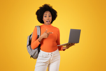 Astonished young African American lady with a backpack points at a laptop screen