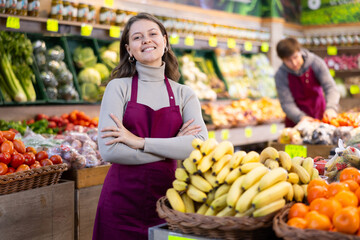 Happy young female seller standing by basket full of bananas in large grocery market