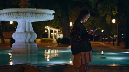 Young woman scrolling mobile phone standing near city fountain late evening.