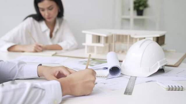 Professional architect engineer drawing blueprint and working together with young beautiful caucasian coworker on meeting table with safety helmet, house model, blueprint scatter around. Immaculate.