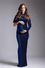 Portrait of young pretty pregnant woman on gray studio background. Female in blue sequin dress with hands near pregnant belly
