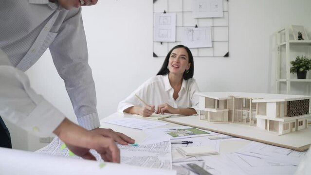 Skilled caucasian interior designer working with house model while discussing with asian architect engineer about house plan on meeting table with blueprint and house model scatter around. Immaculate.