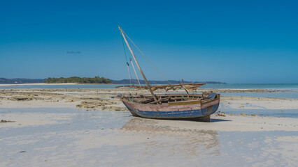 An old weathered fishing boat tilted into shallow water at low tide. A mast, a folded sail against the blue sky. Reflection in a puddle of water. The ocean, a green island in the distance. Madagascar.