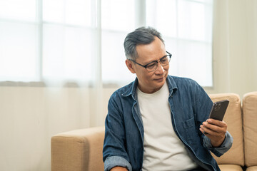 Handsome asian mature old man using smartphone on sofa in living room at home. Happy Portrait of cheerful smiling senior asian man holding cell phone. Mature People and lifestyle