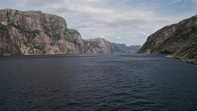 Aerial shot, flying low over the water of Lysefjord, Norway. High, vertical cliffs reach up to the bright, overcast sky on the left, while a rocky, moss covered shoreline passes on the right. 