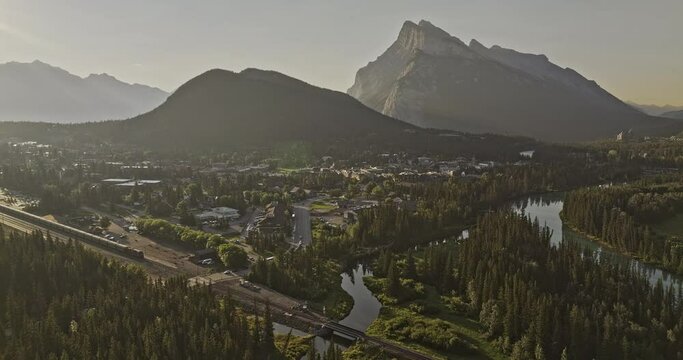 Banff AB Canada Aerial v33 cinematic flyover capturing scenic landscape of train crossing quaint town by the Bow river and Rundle mountain ranges at sunrise - Shot with Mavic 3 Pro Cine - July 2023