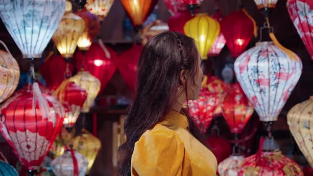 Attractive Vietnamese woman in yellow outfit looks at lantern shop in Hoi An at night