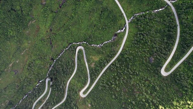 Bird's Eye View, tracking over a winding serpentine road in thick trees and grass. Tight hairpin bends snake up the steep shadowy mountainside. The road crosses a thin, craggy stream. Slow motion.