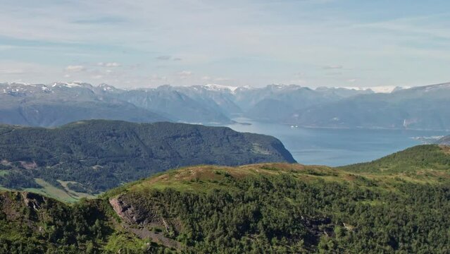 Aerial shot, soaring over a tree lined ridge, in Norway, revealing snow capped mountains and a Fjord in the background. 