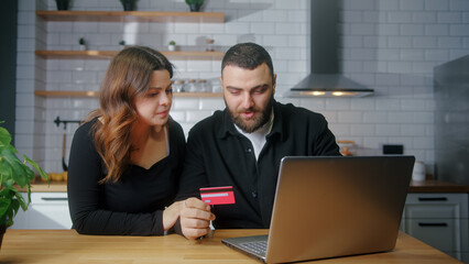 Couple sit in the kitchen picking goods to buy man enter credit card number on laptop for shopping...