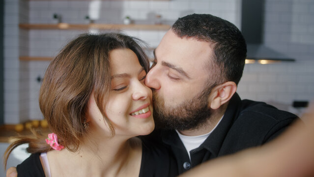 Young couple in love making a selfie using phone having fun in the kitchen looking at camera smiling. Man kiss to girl