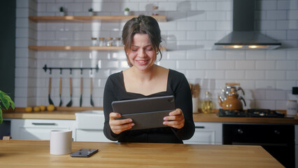 Young woman playing a video game on a tablet computer while sitting in kitchen at home. Excited...