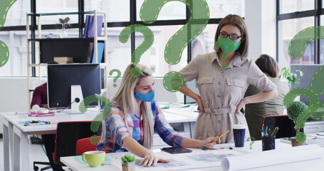 Image of question marks over diverse businesswomen wearing face masks and using computer