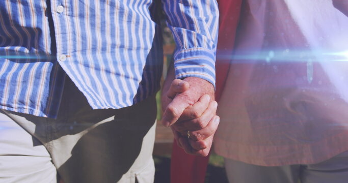 Image of light moving over midsection of senior couple walking in park holding hands
