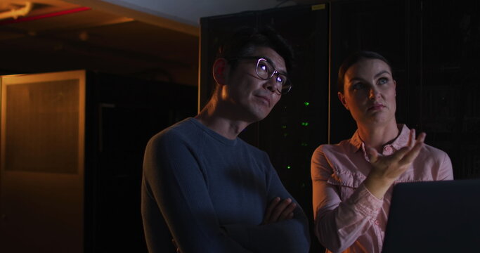 Caucasian woman and Asian man review data in a dim server room
