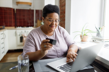 Happy african american plus size senior female in glasses shopping online using laptop and plastic debit or credit card, enjoying special offers and sales on marketplaces, sitting at kitchen table