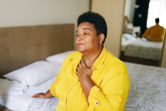 Overweight black elderly female having problems with health touching throat and chest, feeling exhausted. Upset sad depressed african american senior woman grieving sitting on bed in bedroom