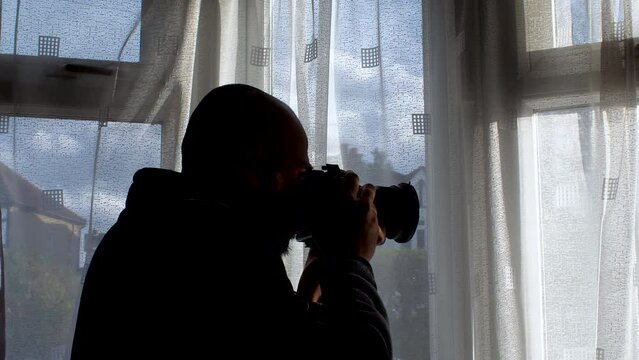 The individual peers through the window, camera in hand, framed by drawn curtains, evoking a sense of detective observation.