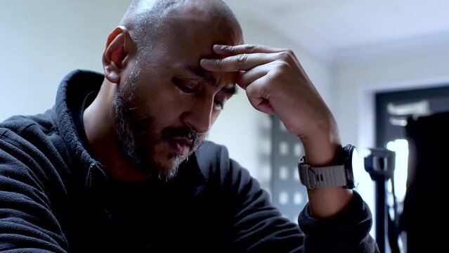 A man sits, his hand scratching his head, his expression revealing the burden of stress and anxiety he carries, vividly illustrating the tangible strain he experiences.