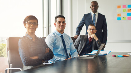 Four diverse successful business partners sit in a conference room, in an office building and look confidently into the camera, while there are notes and a laptop on the table - 756994264