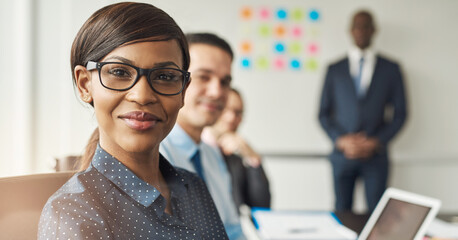 A young smiling black businesswoman sits at a conference table with her colleagues in the background. She looks confidently and happily into the camera - 756994267