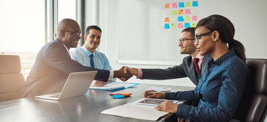 Two diverse business teams sit at a conference table and shake hands on reaching an agreement. They all smile at having made a business deal