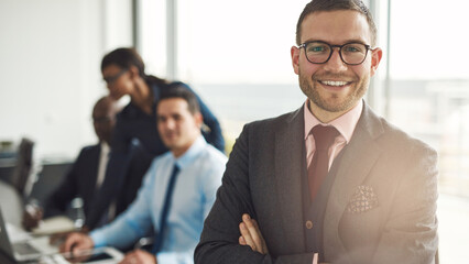 Confident handsome young businessman stands in a boardroom, surrounded by diverse colleagues. He has his arms crossed and looks directly into the camera with a big smile - 756994459