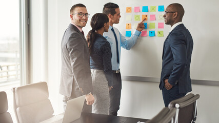 Group of diverse business people. The CEO points to a sticky note on a whiteboard while standing with colleagues in the office boardroom. A businessman looks back over his shoulder and smiles - 756994624