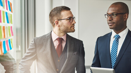 Two focused and serious diverse males executives standing in an office boardroom behind a glass wall with sticky notes in a discussion - 756994667