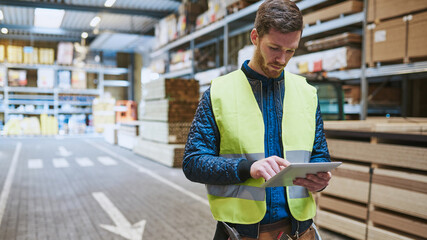 Young warehouse worker consulting a tablet while wearing a yellow safety vest