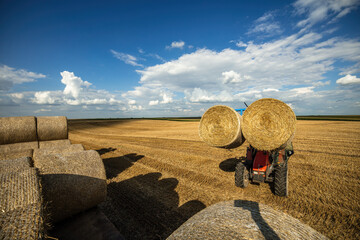 Tractor gathers large round hay bales under a vast, blue sky atop the golden field