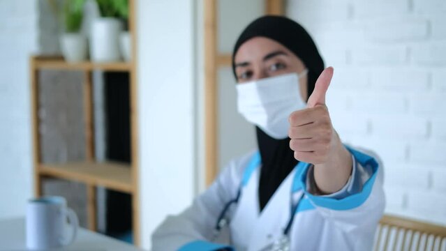Muslim woman doctor shows thumbs up, side view. Selective focus: Muslim woman doctor in medical gown, mask and black hijab shows thumbs up. Concept of thumbs up gesture as approval and agreement