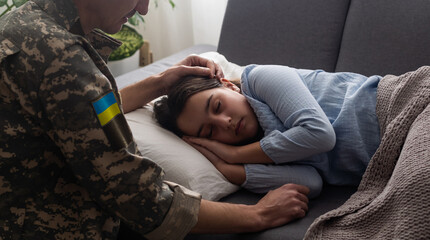 Soldier in Ukrainian military uniform kissing his daughter while she sleeping on sofa at home....