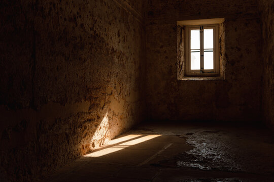 Old empty room with stone walls, almost in darkness, with the only light from the window that can be seen as it is projected on the floor and the wall.