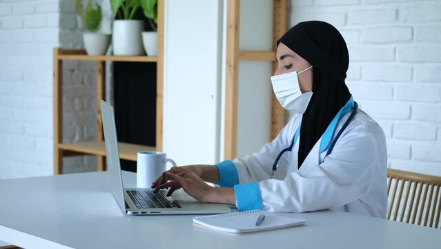 Muslim woman doctor typing on laptop. Close-up shot: Muslim woman doctor moves laptop and works on keyboard, writing result in notebook. Concept: laptop helps doctor to keep documentation