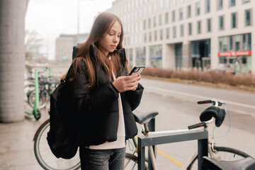 School age girl renting an electric scooter or bicycle using smartphone, making contactless payment through mobile app. Bicycle rental in the city in the autumn-winter season - obrazy, fototapety, plakaty