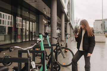 School age girl renting an electric scooter or bicycle using smartphone, making contactless payment through mobile app. Bicycle rental in the city in the autumn-winter season - 757021066